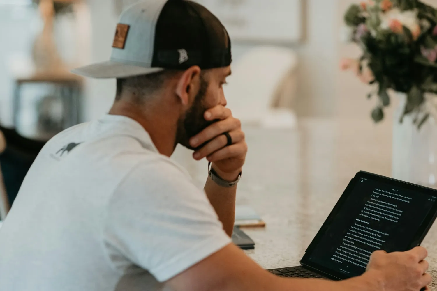 A person in a cap reading a document on a tablet at a kitchen counter, one hand resting against their chin.