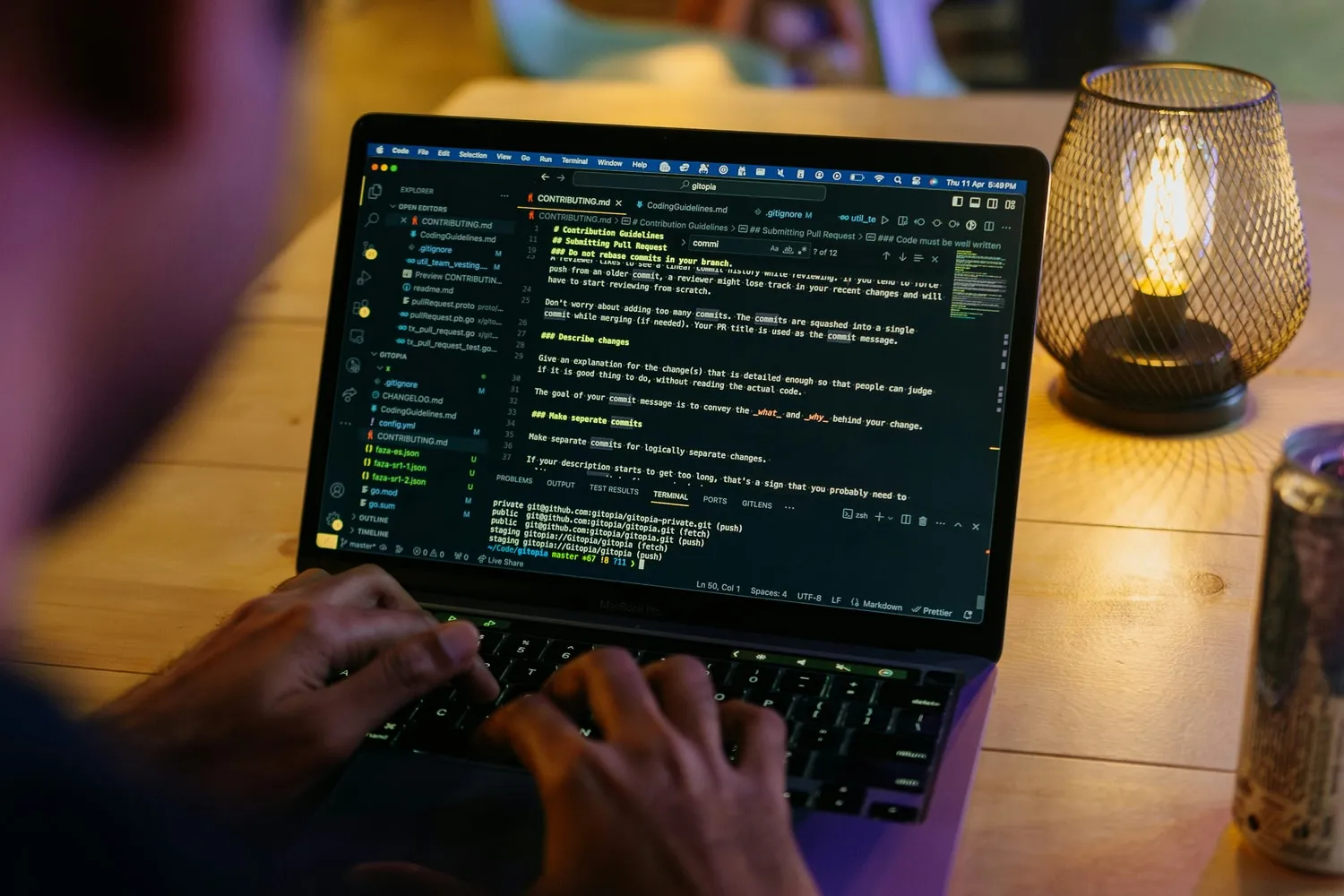Hands typing on a laptop at a wooden table, a code editor open on screen, with a warm lamp beside it.